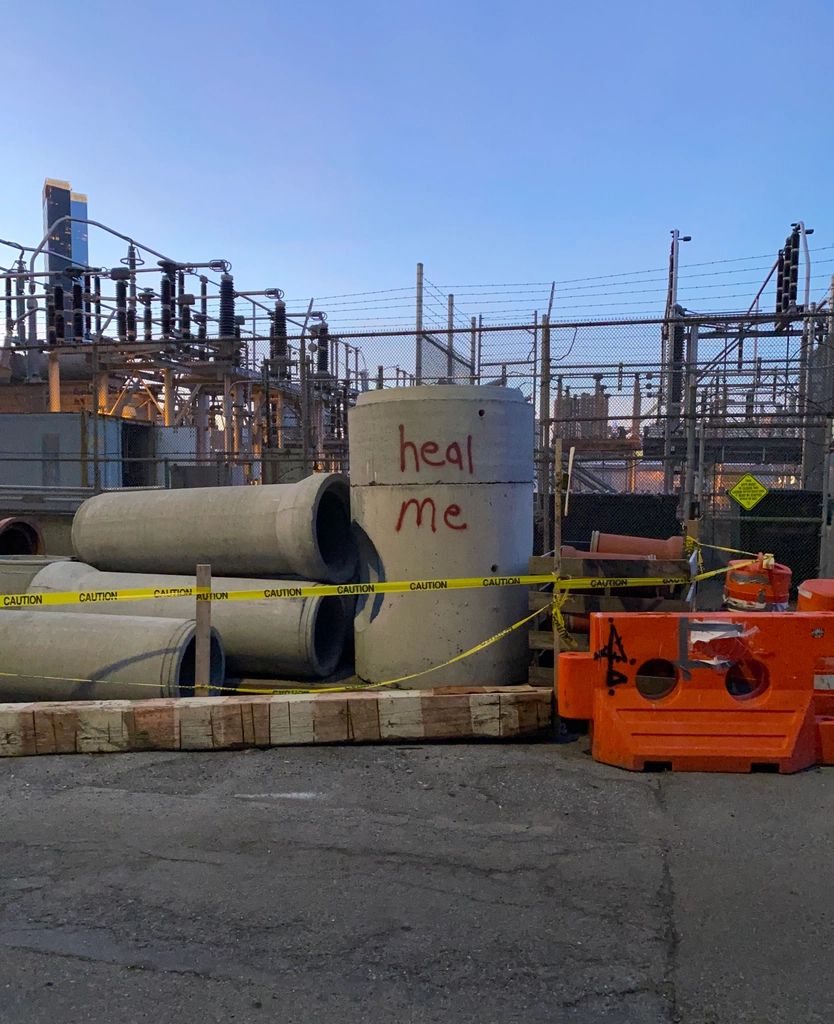 Large concrete tubing, fenced off with yellow caution tape, is piled outside an electric substation. There is an ethereal glow from the substation's lights and the almost-dusky sky. "heal me" is graffiti-ed on the largest, upright tube.