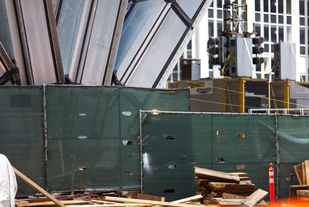 A construction site at the base of a large building is littered with loose planks. There's some out-of-focus electrical machinery in the background.