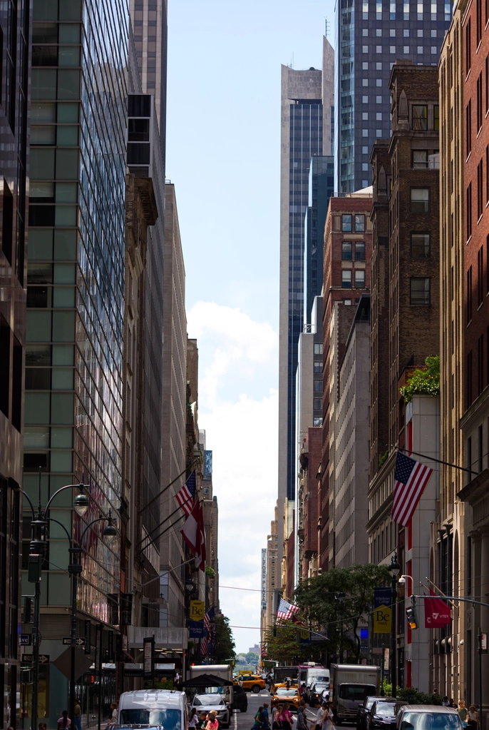 Rows upon rows of skyscrapers line an avenue bustling with activity as far as the eye can see. Light bounces back and forth between the glass-coated buildings. There are many american flags hung up.