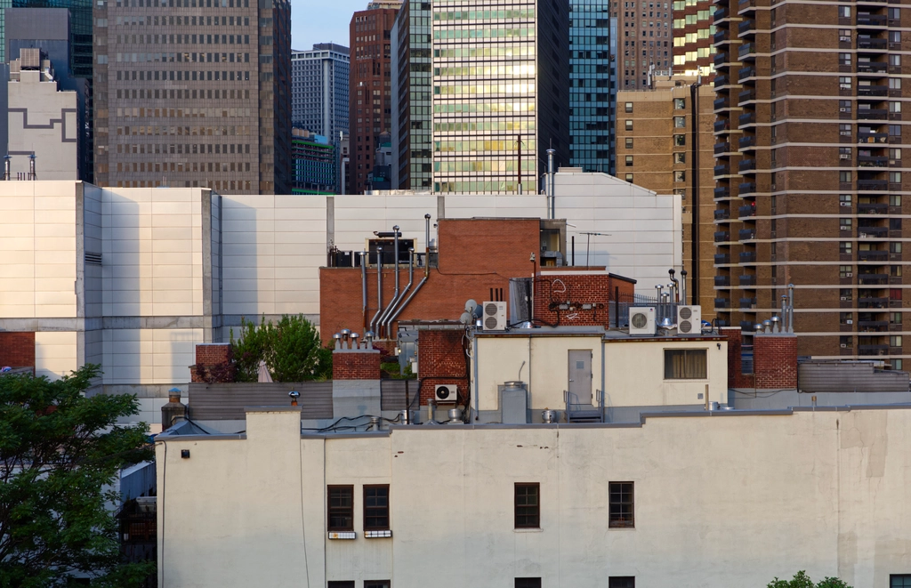 A small rooftop area containing a small garden and plenty of AC units, sits tilted in front of a sound wall, in front of myriad skyscrapers, all at slightly different angles. The morning sunlight gives things a peaceful look.