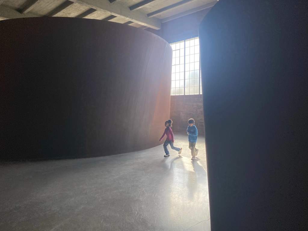 Two children are playing between two giant rusted elliptical metal structures that reach almost the entire way to the wooden roof of the brick building they're in. Hazy sunlight from the window behind them leads to splotchy gradients in the darker areas.