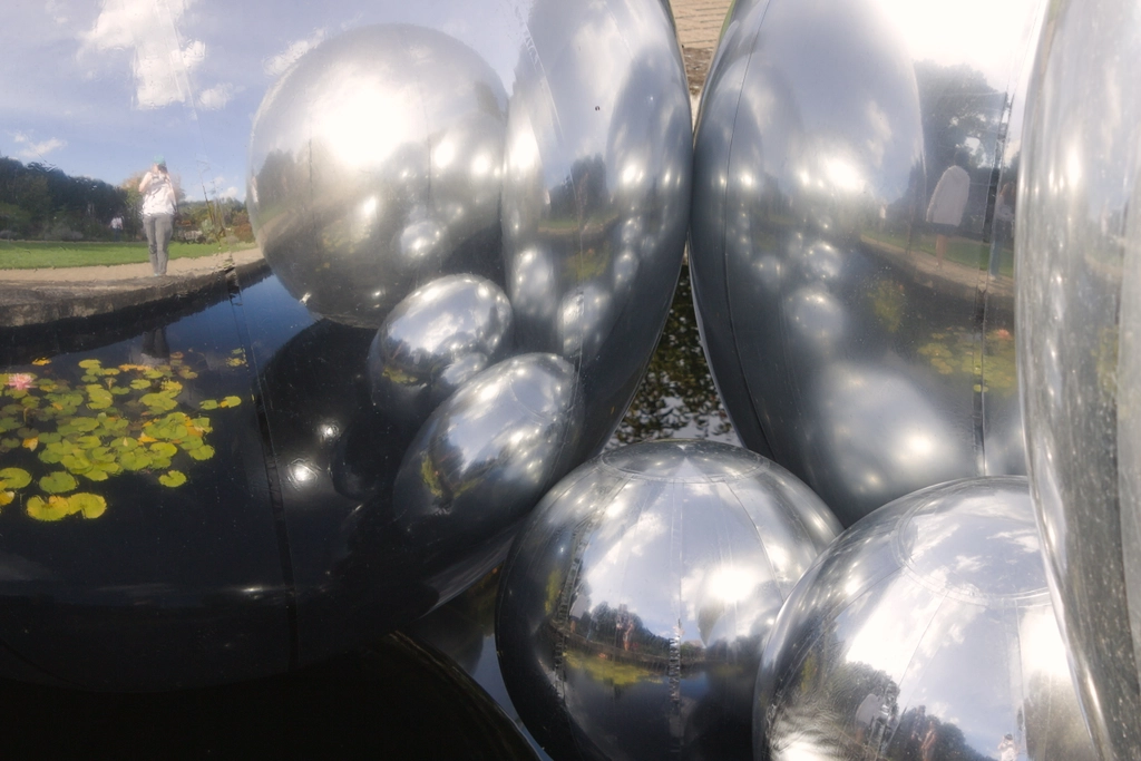 The photographer is partially reflected in the larger part of a reflective ball sculpture floating in the middle of a small man-made pond. The myriad balls of different sizes reflect each other through a thin layer of dust that diffuses their highlights.