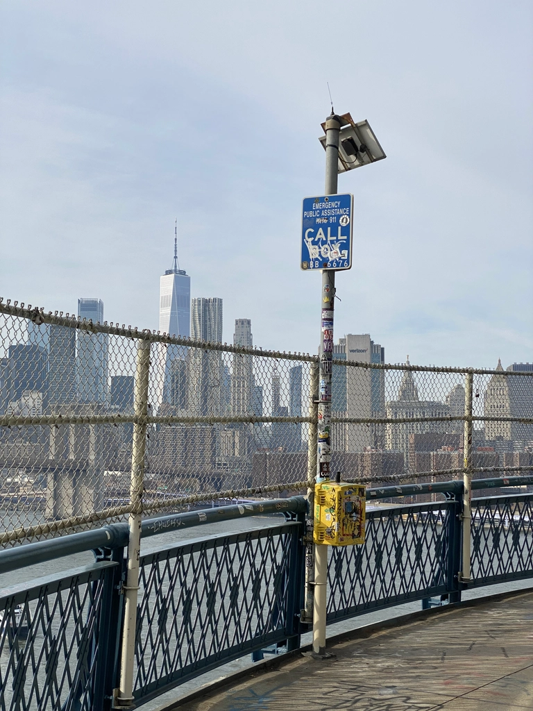 An emergency call box is plastered with all manner of stickers and graffiti, overlooking WTC One from the middle of the East River.