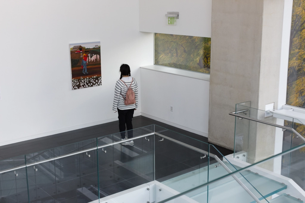 A person stands a respectful distance away from a painting, looking intently. There are no other paintings on the walls in this upscale stairwell. Fall trees blow in the wind outside the windows.