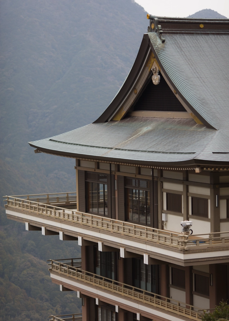 A white-yellow-brown-gray shrine building with three levels of balconies overlooks some hazy mountains.