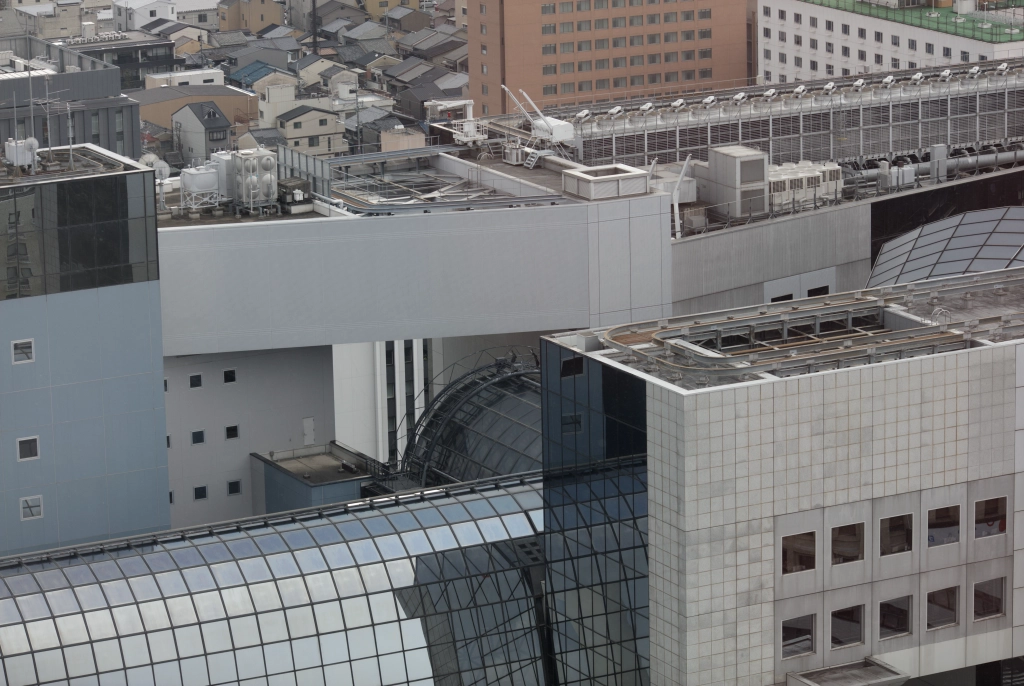 A long series of impossibly-connected buildings separates a glass canopy from rows of houses & apartments in the background.