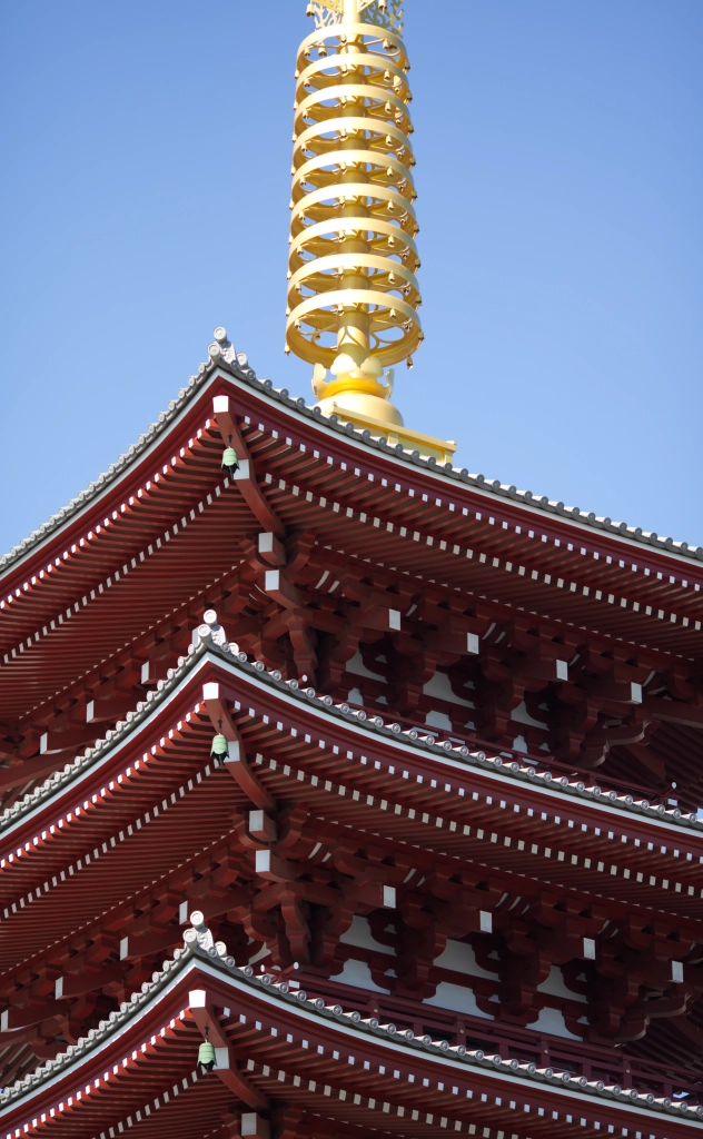 Three stories of a red pagoda & that golden ringlike buddhist spire thingy are viewed from below. The blue sky is perfectly clear.