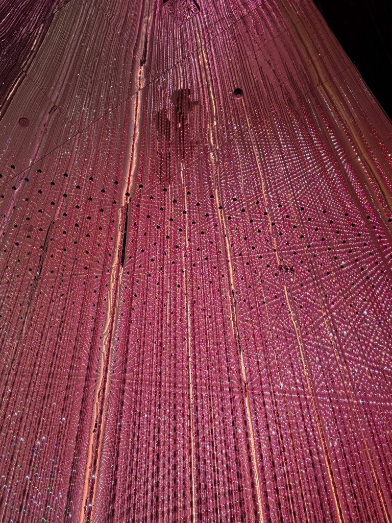 Strands of red-ping fairy lights are suspended from the ceiling in neat rows, forming further lines & intersections at all the different vanishing points. Some people & sensors can be seen in the mirrored ceiling's reflection.