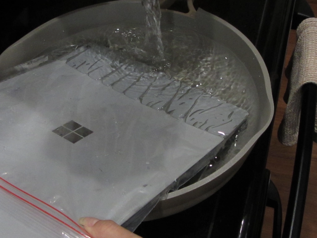 A laptop in a plastic bag with one edge submerged in a large sauce pan, with steaming water being poured over that edge.