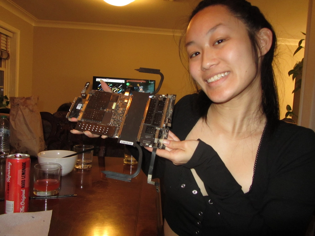 Holly holds up a laptop motherboard in a connected dining/living room. Dishes from the group dinner are visible on the table, and the TV screen shows a video game being played in the background.