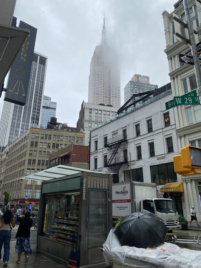 The Empire State Building is partially shrouded in a small low cloud, behind an old storefront, a city-grade Newsstand, and "Swingers: The crazy golf club".