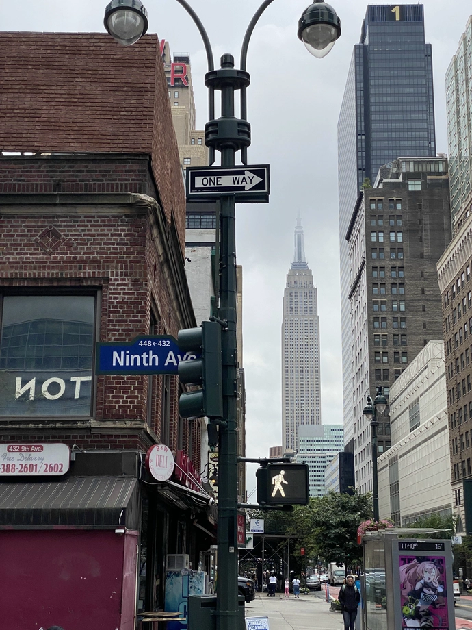 The Empire State Building is a straight shot down the street, behind roadside trees, an old brick pizza building, an anime game ad, and a walk sign.