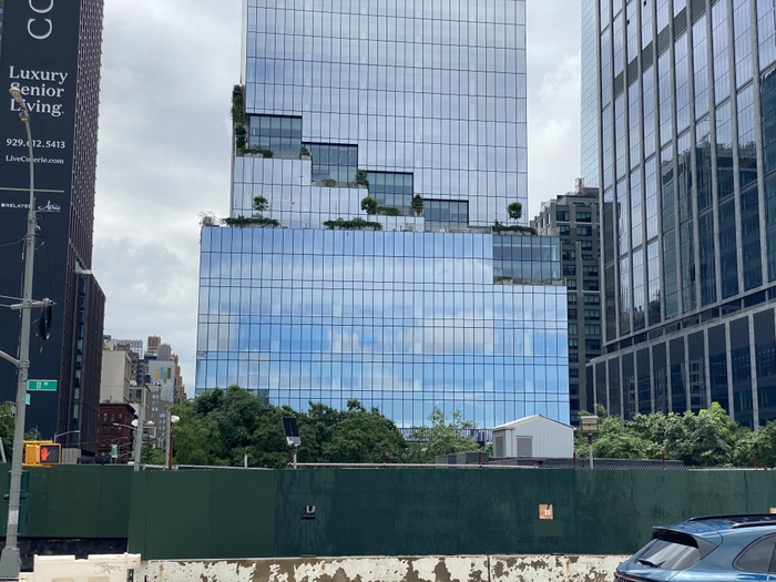 A new-looking stair-steppy skyscraper reflects a blue sky against an overcast background, with a green construction lot in front of it. The Empire State Building is nowhere to be seen.