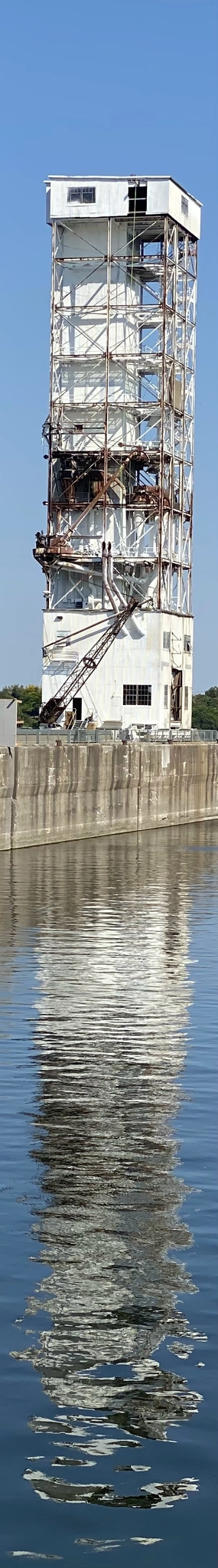 A white, rusting tower, mostly made of empty space between steel crossbeams & piping, is reflected over the concrete pier it sits on to the smooth, barely wavy water