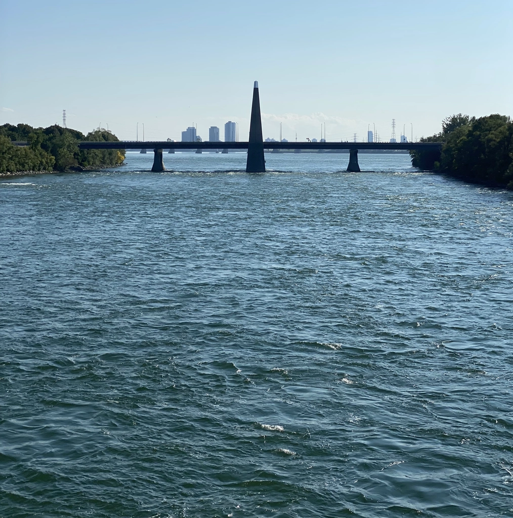 A perfectly symmetrical black bridge, with a tall spire in the middle and two smaller supports on either side, connects two islands over a uniformly choppy dark cyan river.