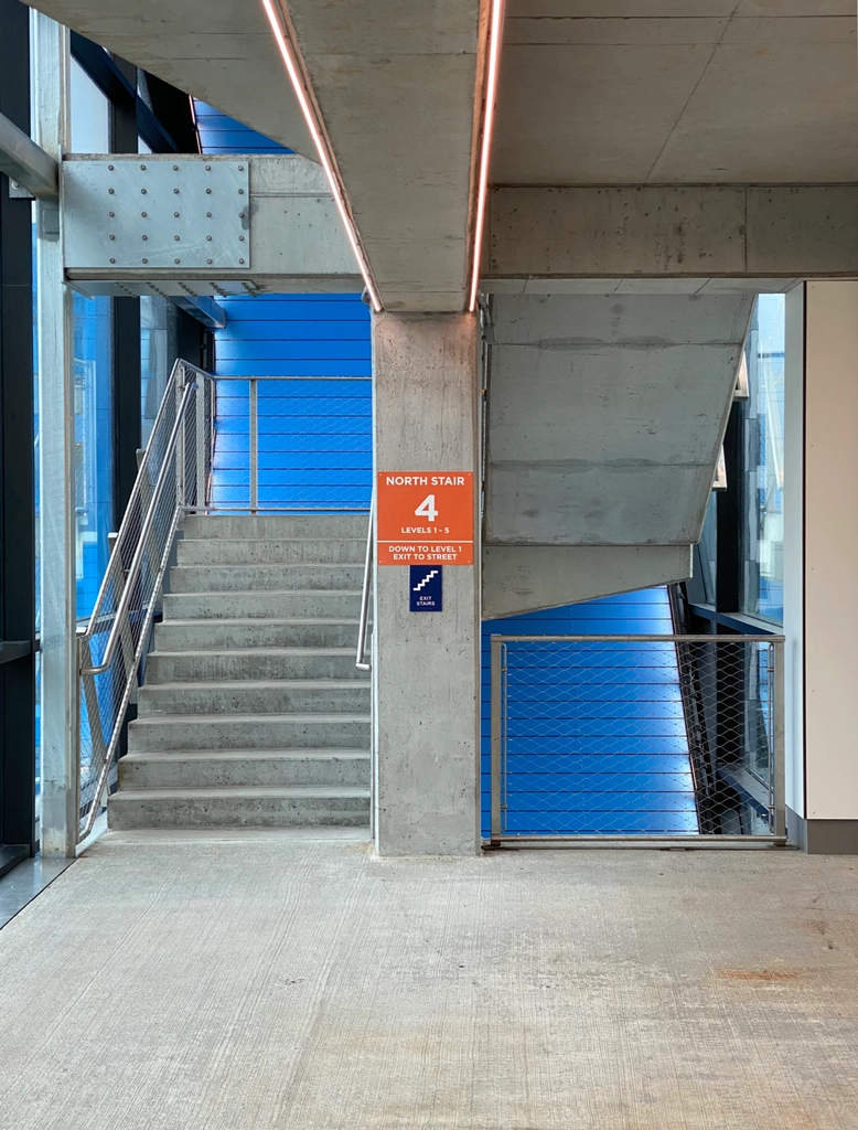 A concrete parking garage stairwell with blue facade visible in the background, and an orange sign reading "North Stair 4", illuminated by soft orange-white light.