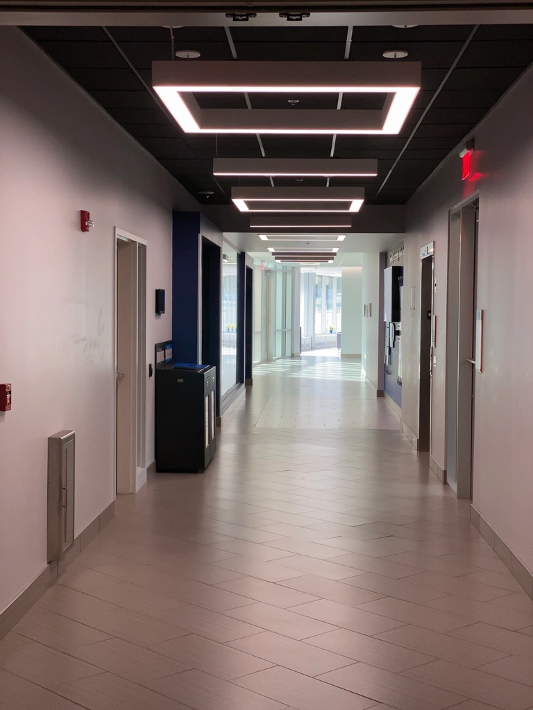 A hallway lit by square overhead lightfixtures close, then natural sunlight through green/cyan-tinted glass further on. A rectangular motif is present throughout.