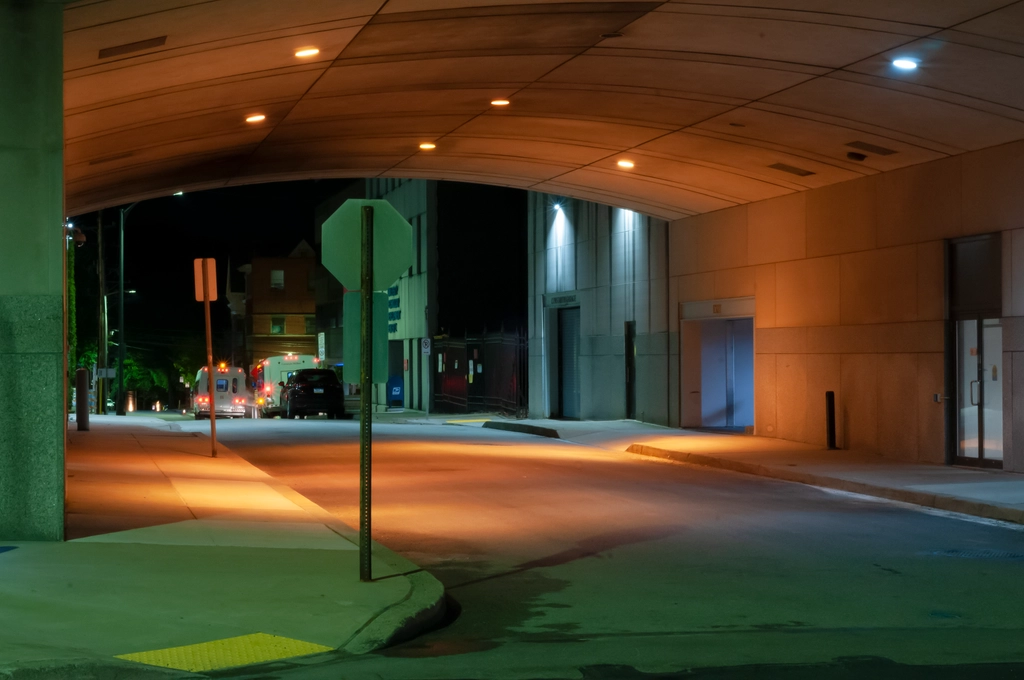 A passageway for cars to go into a garage underneath a building is bathed in multiple colors of light; green from the fluorescent ones outside, orange from the sodium ones in the tunnel, and purple from the lights inside the garage. There are also white lights on a building and red tail lights in the background.