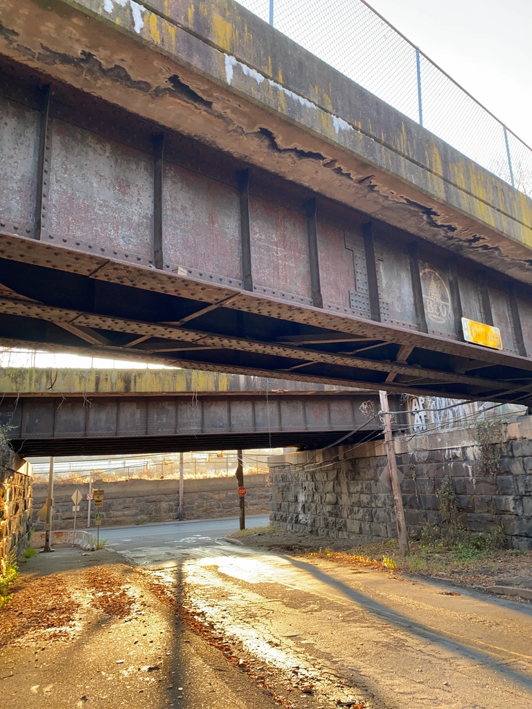 Late afternoon sun streams beneath a rusty bridge, lighting up the pavement and casting long shadows from wooden electrical poles.