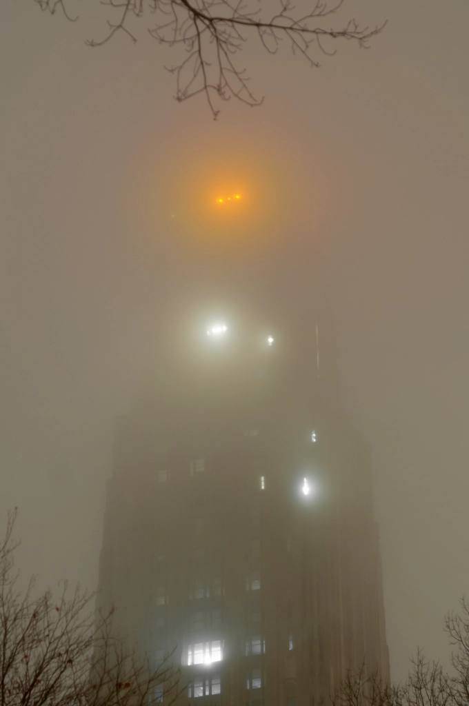 The Cathedral of Learning in Pittsburgh looms eerily through the mist, with winter tree branches framing the scene.