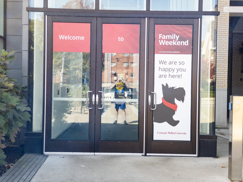 I stand reflected in front of a university building whose glass doors are decorated with a banner "Welcome to Family Weekend!". I am standing alone at the edge of the shadow cast by the building and the whole view is slightly askew.