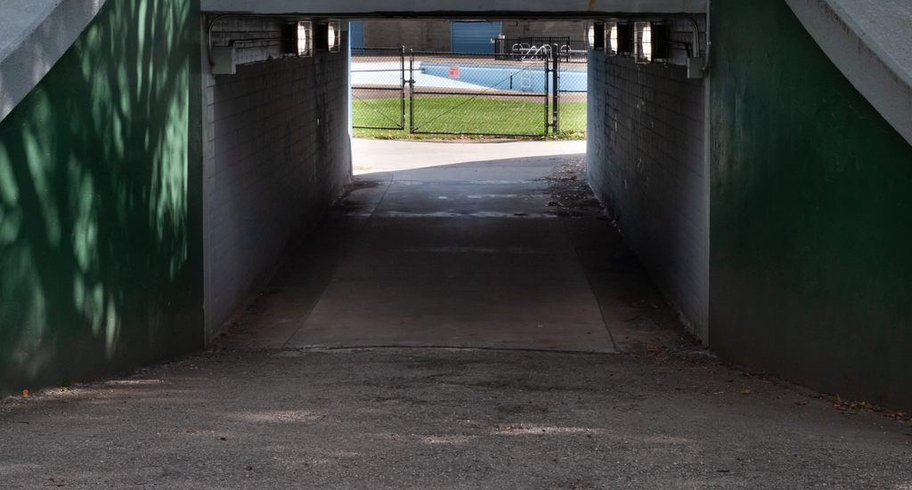 Underpass illuminated by fluorescent lights in the middle of the day. An empty pool is visible though the tunnel.