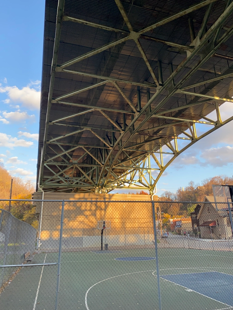 Golden hour sun illuminates a wall by a basketball court underneath a bridge, with trees and tree shadows present. The overall atmosphere is soft.