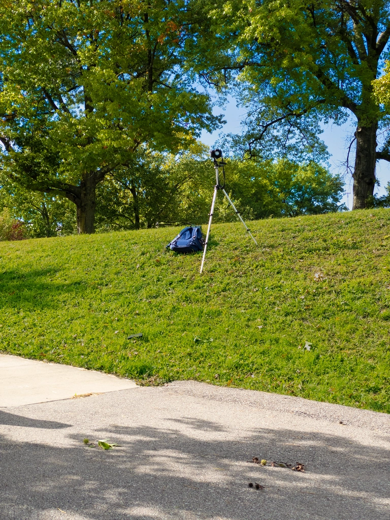 A tripod with a camera rests on a slanted hill with a blue backpack beneath it. The sky and trees and grass are vibrant.