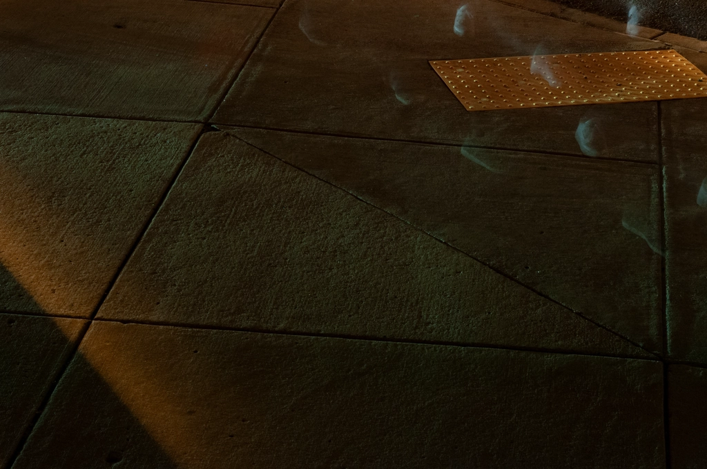 Sidewalk at night is illuminated by headlights from nearby cars. Ghostly footsteps can be seen travelling diagonally across the upper right corner.