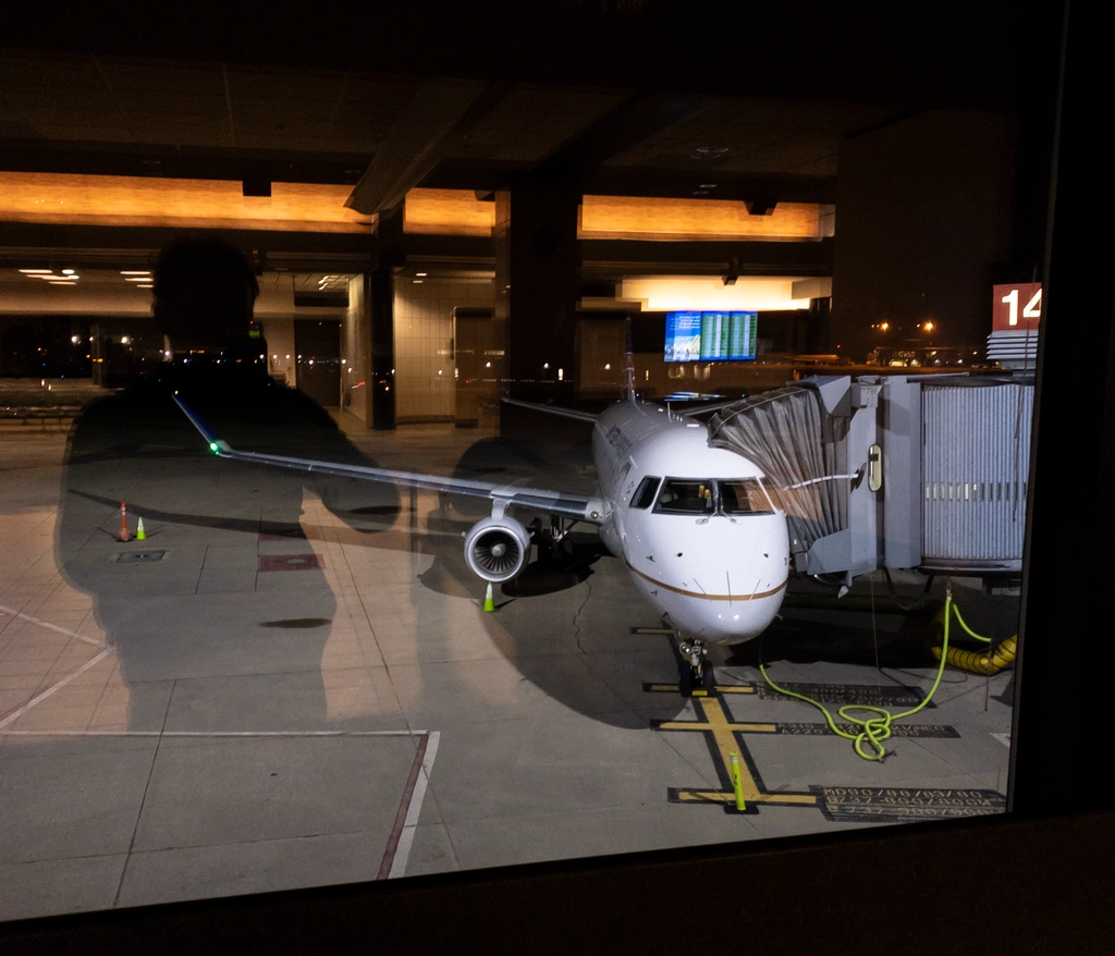 At an airport in the early morning, a plane getting ready on the tarmac is viewed through a window where the reflection of the inside of the airport can be seen, along with the outline of the photographer.