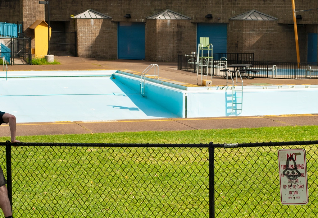 Empty pool from before, now fully visible above a black chain link fence. On the right of the fence there is a vandalized "no trespassing" sign, and on the left there is someone's arm and leg cropped off scaling the fence.