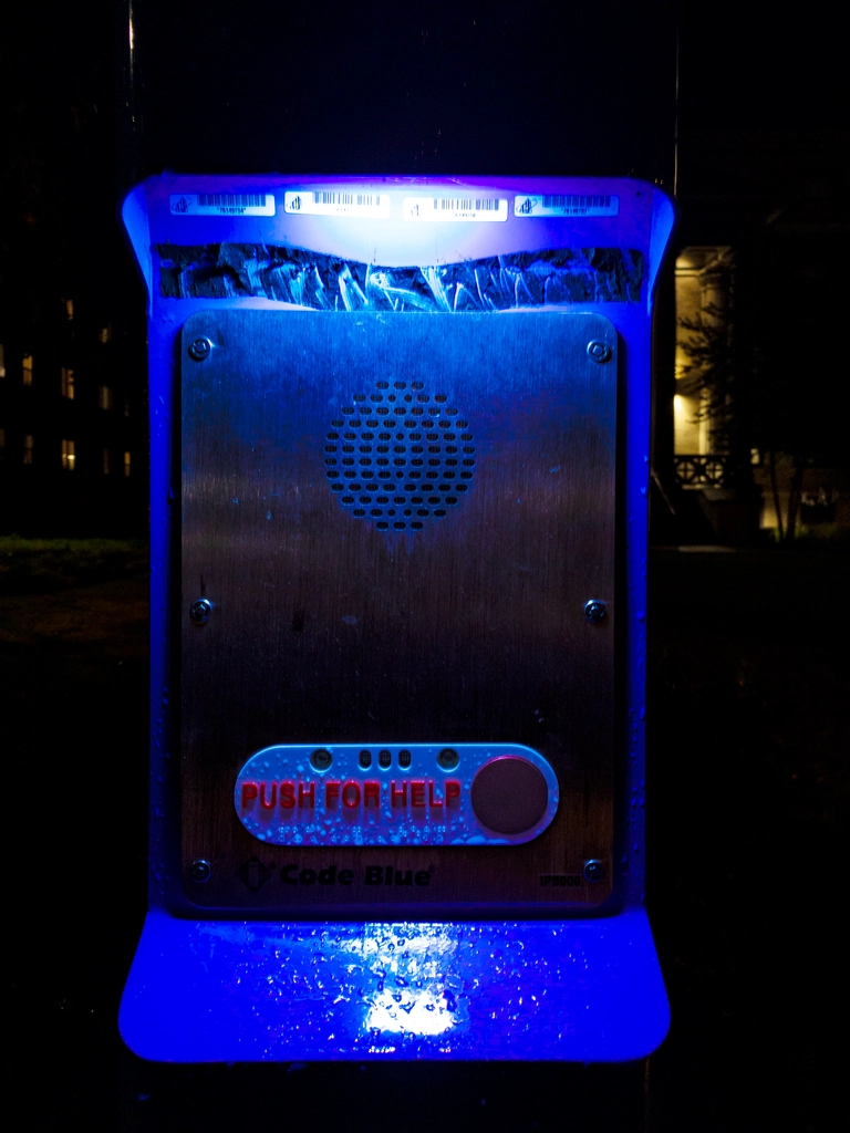 An emergency blue light post at night. It is a close-up shot of the lower blue light illuminating a red "push for help" button