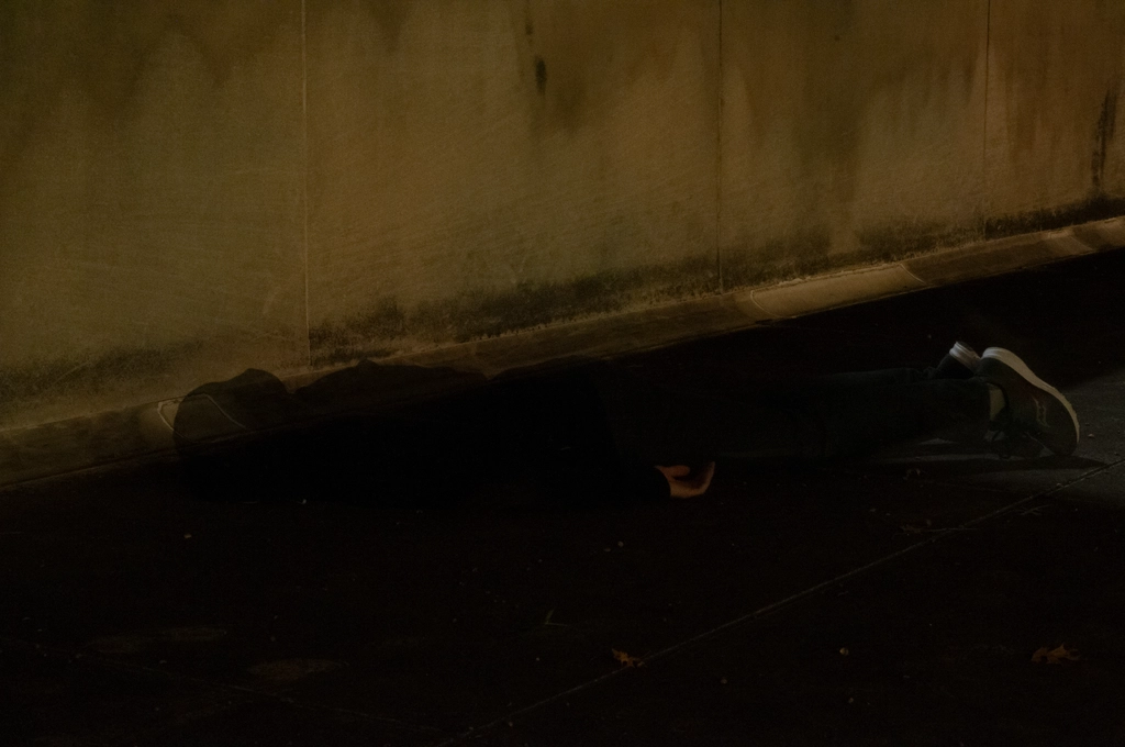 A ghostly figure lies face-down next to a yellow concrete wall at night.