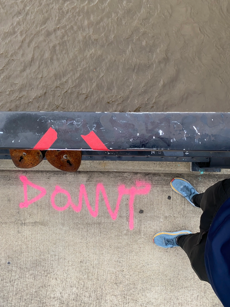 Looking down directly over the railing of a bridge, with the top half of the image brown water and the bottom half concrete with a pair of legs standing on it. There is pink graffiti saying "DONNT" in front of where the legs are standing.