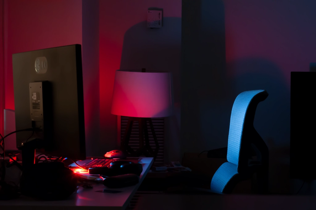 A laptop on a desk glows red from keyboard LEDs, illuminating the surrounding walls and turned-off lamp, while the display shines blue light on an empty chair