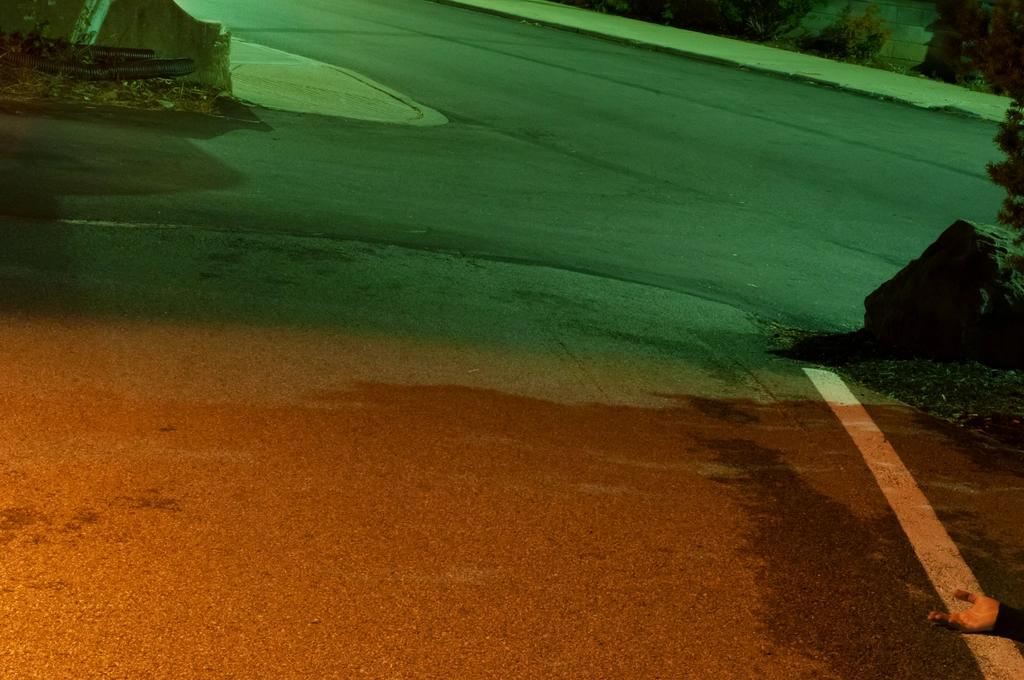 Mixed light of a green fluorescent bulb and an orange sodium lamp create distinct shapes on the ground in a slope leading out of a parking lot. In the bottom corner, a hand lies face-up (cropped off)