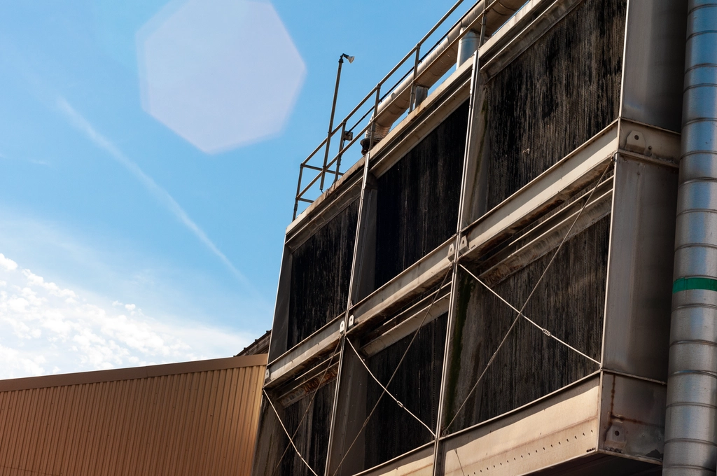 Large air intakes for a power plant reflect light onto a nearby building. The sky is a clean blue and there is a single lens flare from a sun out-of-frame.