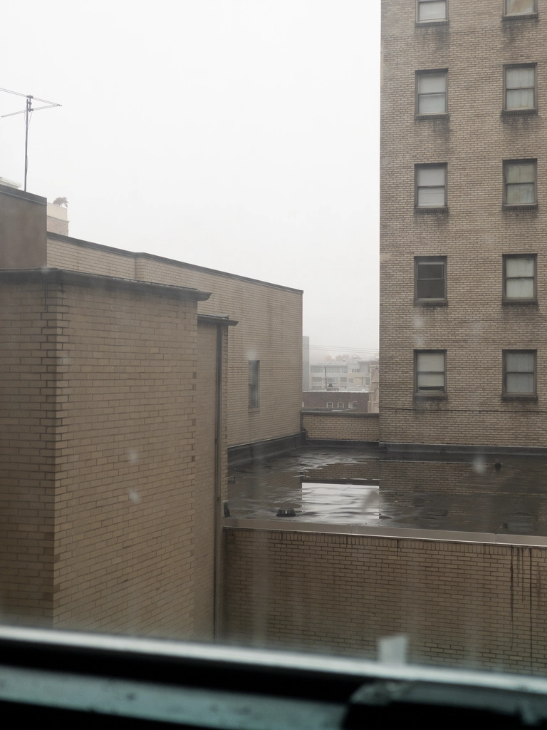 A picture through the window of an apartment building of the other towers. It is currently raining and the lighting from the overcast sky at midday is very drab and depressing.