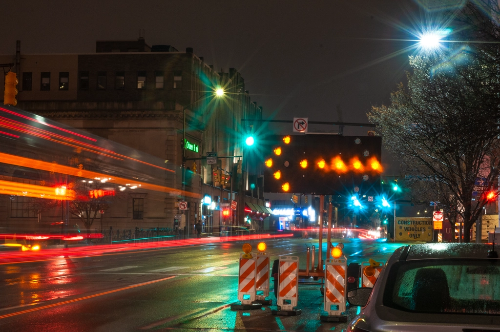 Travelling cars and buses leave streaks of red tail lights on the night road. Small city lights illuminate the wet road and bright construction markers. There is also a big arrow redirecting traffic.