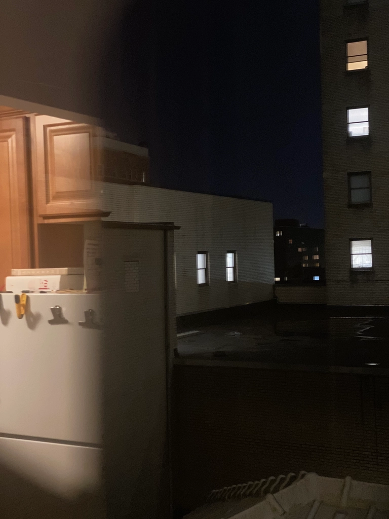 Photo taken at night out the window of an apartment building, viewing the other towers of the building. Part of the outside view is obscured by the reflection of the kitchen in the window.