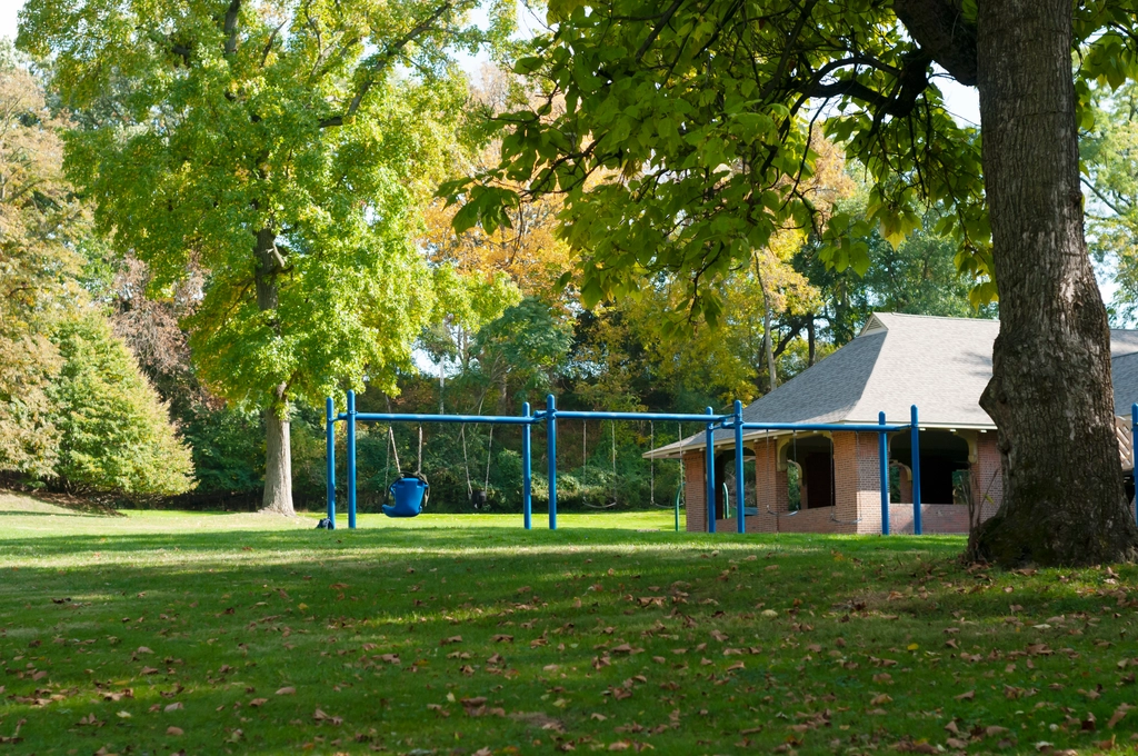 A blue swingset next to an outdoor pavilion in the middle of a lush green grassy field surrounded by fall trees. An empty jacket and backpack are on the child swing.