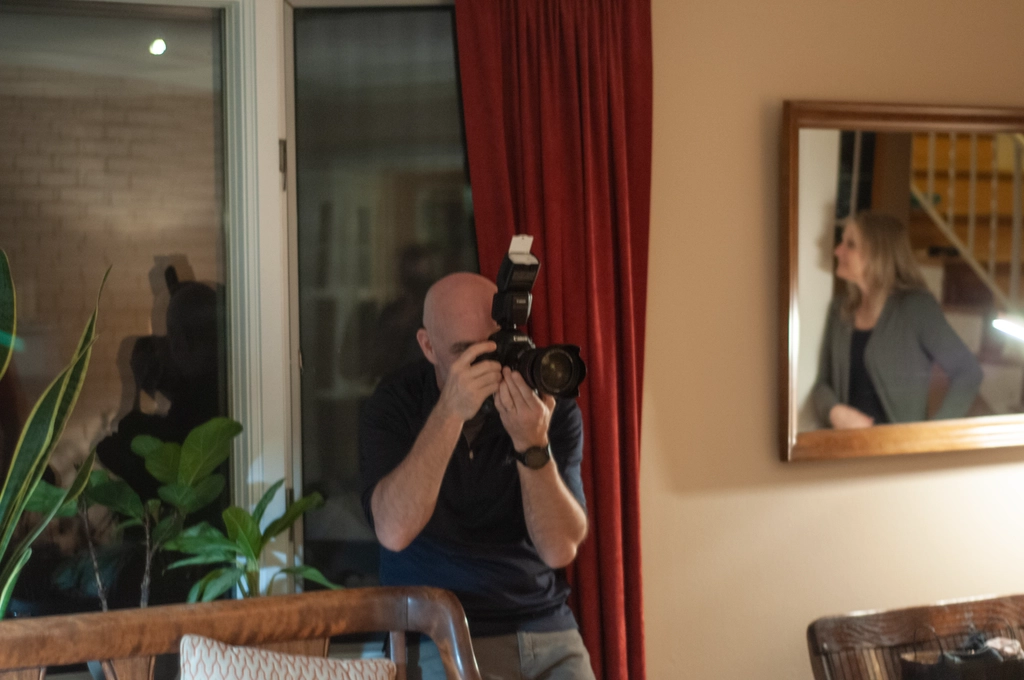 A bald photographer pushes themselves into a corner to get the right angle for an indoor portrait photo of subjects off-screen. Another non-photographed lady can be seen in the mirror next to the man. There is a red curtain and green plants and dark windows and brown wood chairs to add variety to the scene.