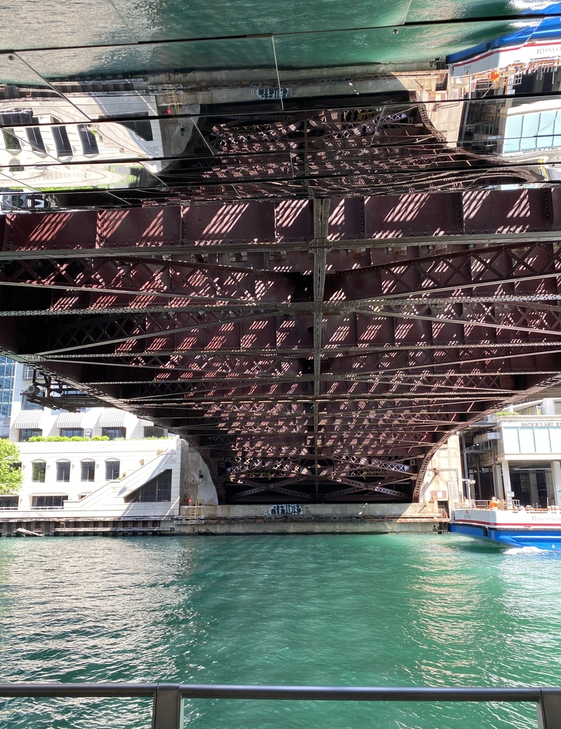 Morning light illuminates the water under a brown-painted metal bridge, as viewed from the riverfront directly underneath.