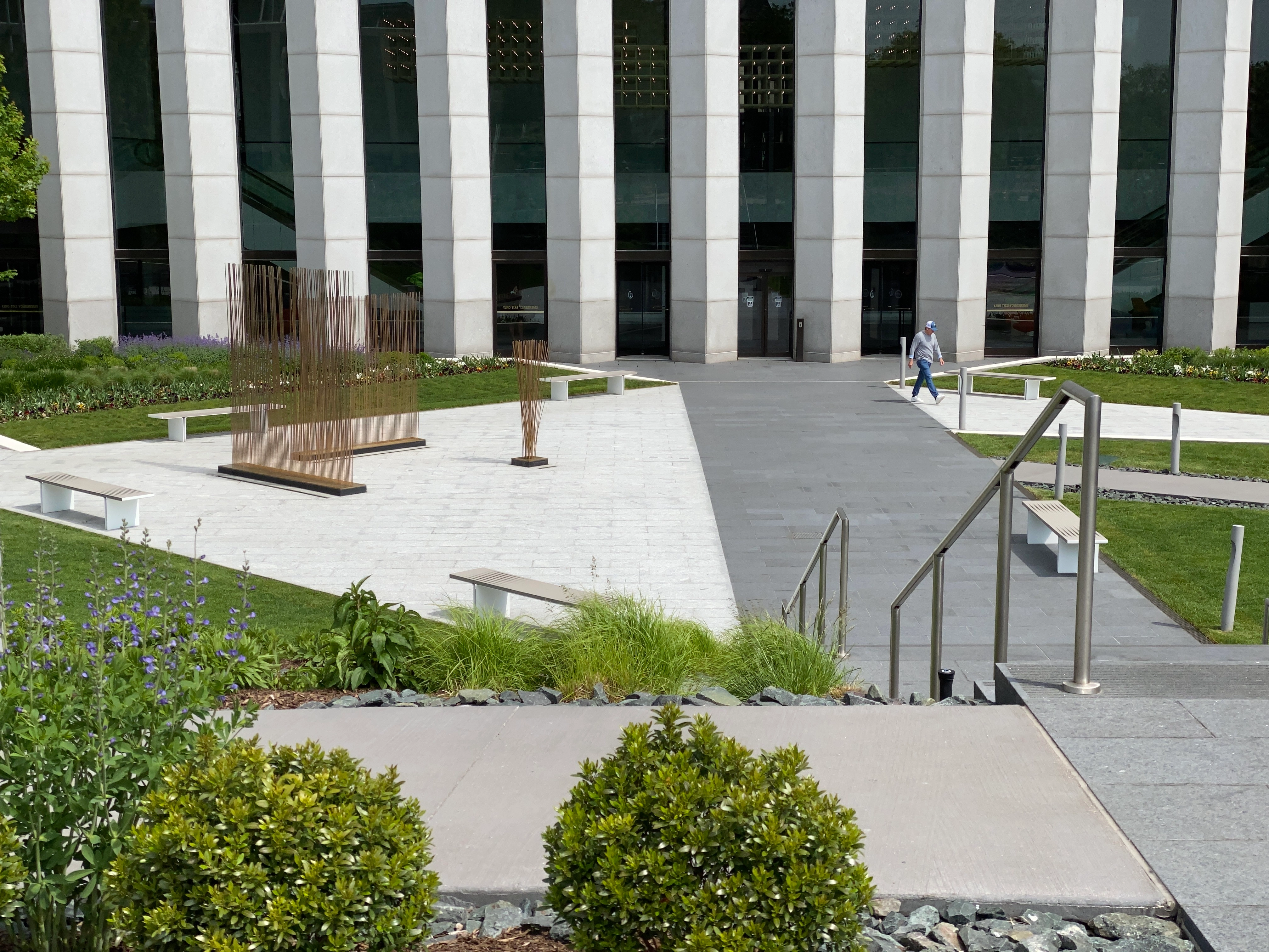 A modern-looking courtyard in front of a building is populated by shrubs, grass, railings, abstract sculptures, and a singular office worker walking awkwardly.