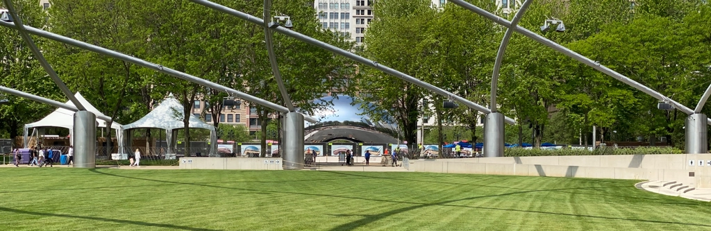 The Bean, with its patio fenced off under construction, as viewed from across the lawn, with a few people milling about.