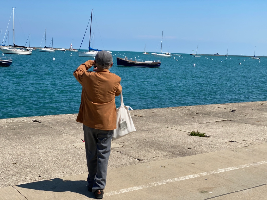 A man in a brown fuzzy jacket takes a picture of the sailboats on Lake Michican with his phone, his beret tangent to where the dark blue of the water becomes the light blue of sky.