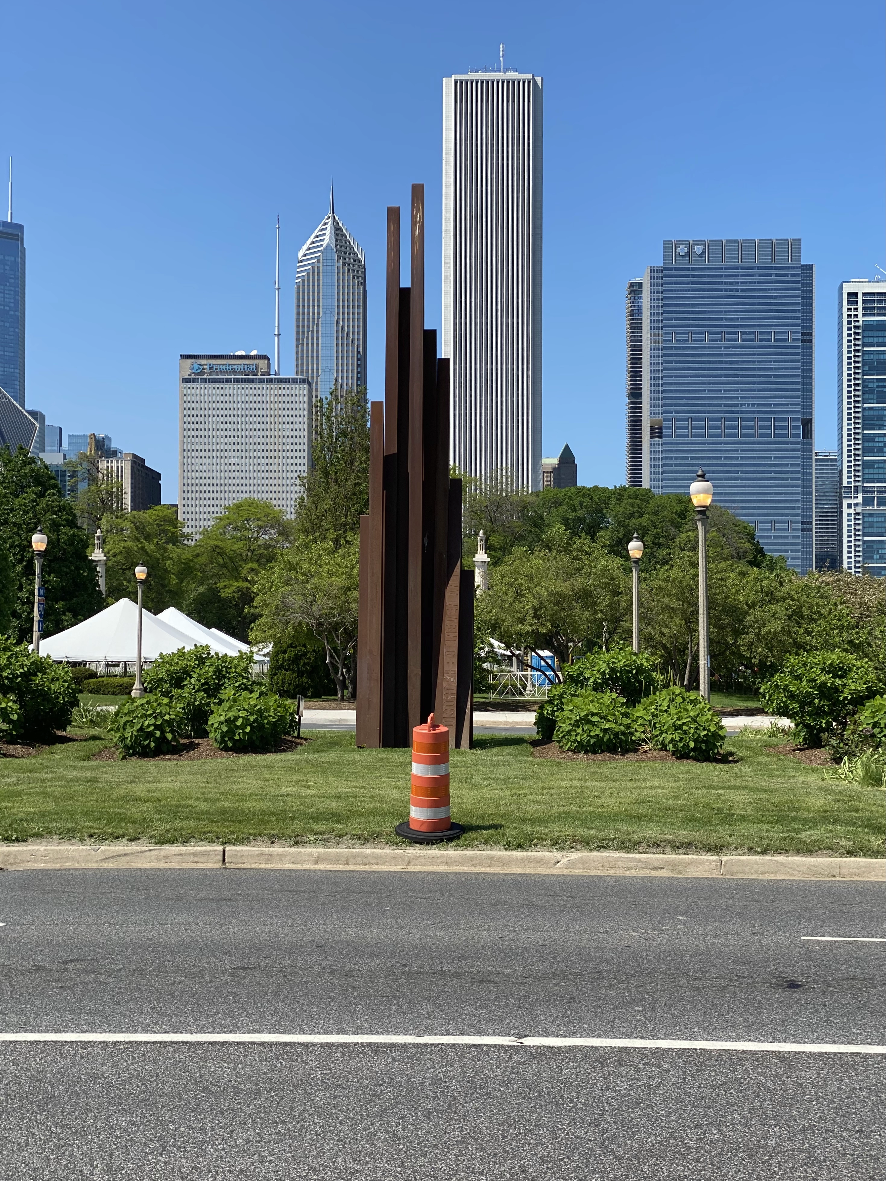 An orange traffic cone sits on a grassy median in front of a (purposefully?) rusted sculpture in front of a Northern Downtown Chicago skyline. Everything lines up.