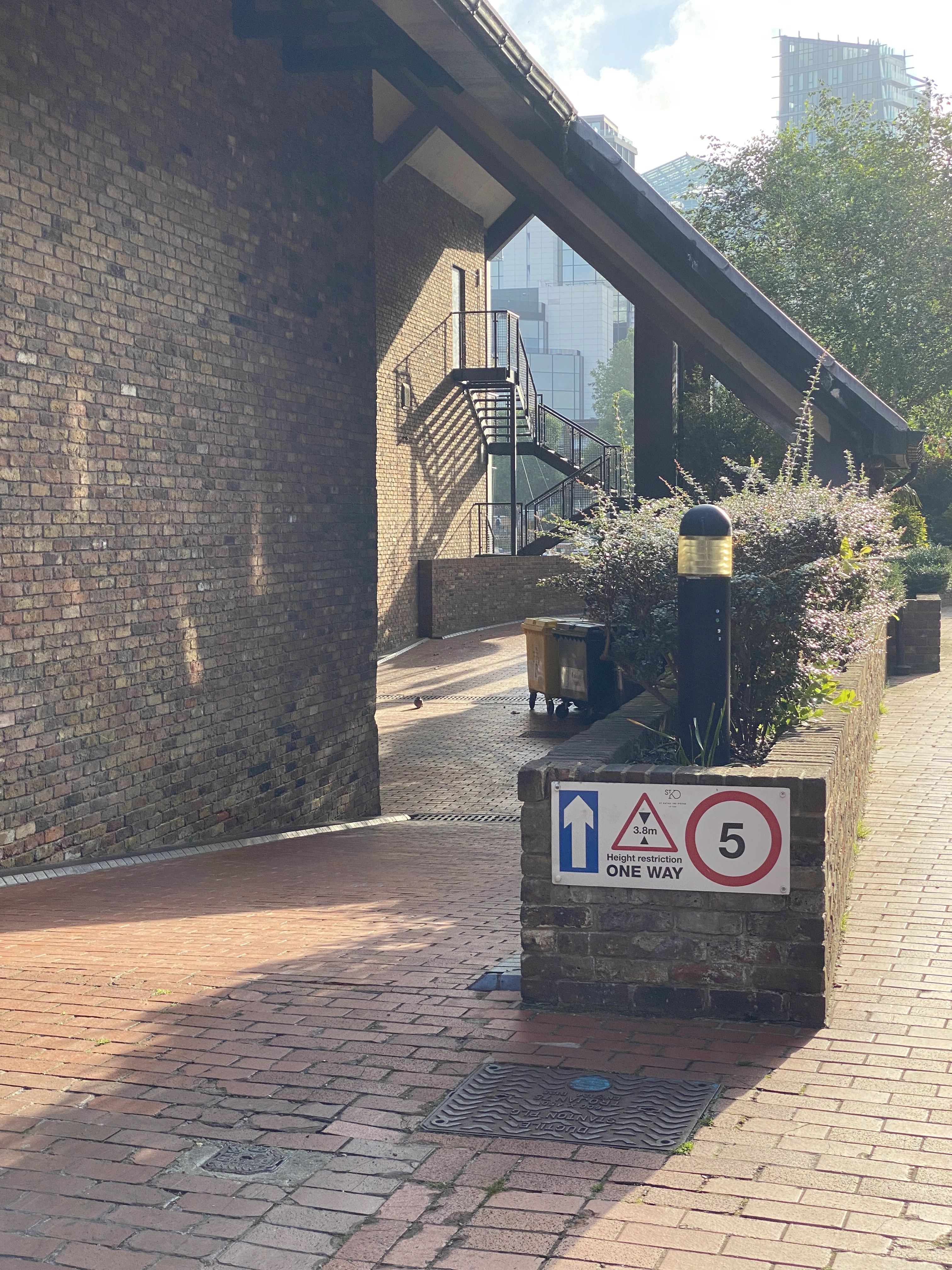 Sunlight streams onto a red brick walkway, with some shrubbery and a lamppost in the foreground leading to a fire escape stair in the background. There is some signage posted.