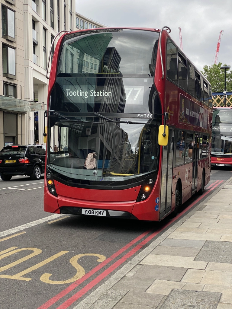 A double-decker red bus labeled "Tooting Station 77" approaches the camera, with the city reflected in its full-frontal windshield. There is a like bus behind it.