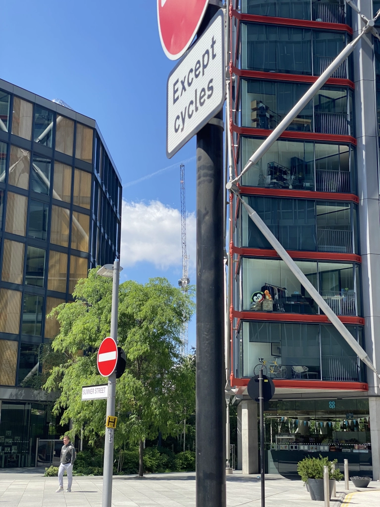 Modern apartment buildings with external structural X-supports and floor-to-ceiling glass windows surround a courtyard where one person is standing in a hoodie. A road exit is labeled with a "Do Not Enter" symbol.